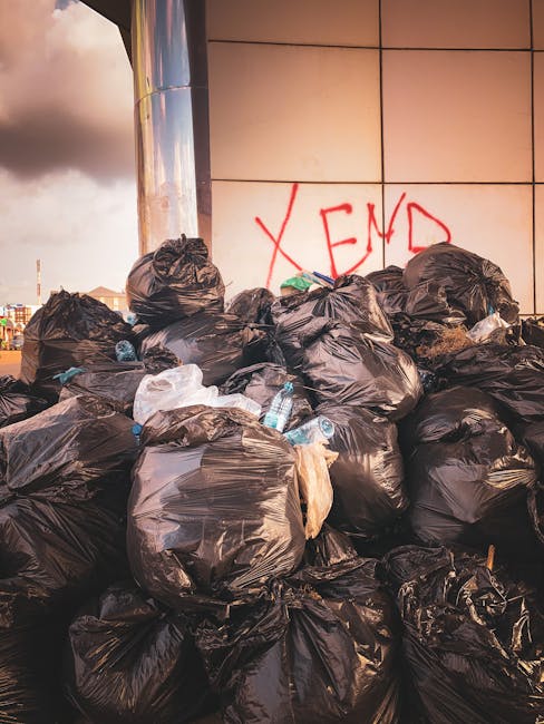 A large pile of black garbage bags filled with waste, stacked against a wall with graffiti that reads 'XEND' in red spray paint. Several plastic water bottles and miscellaneous trash items are visible