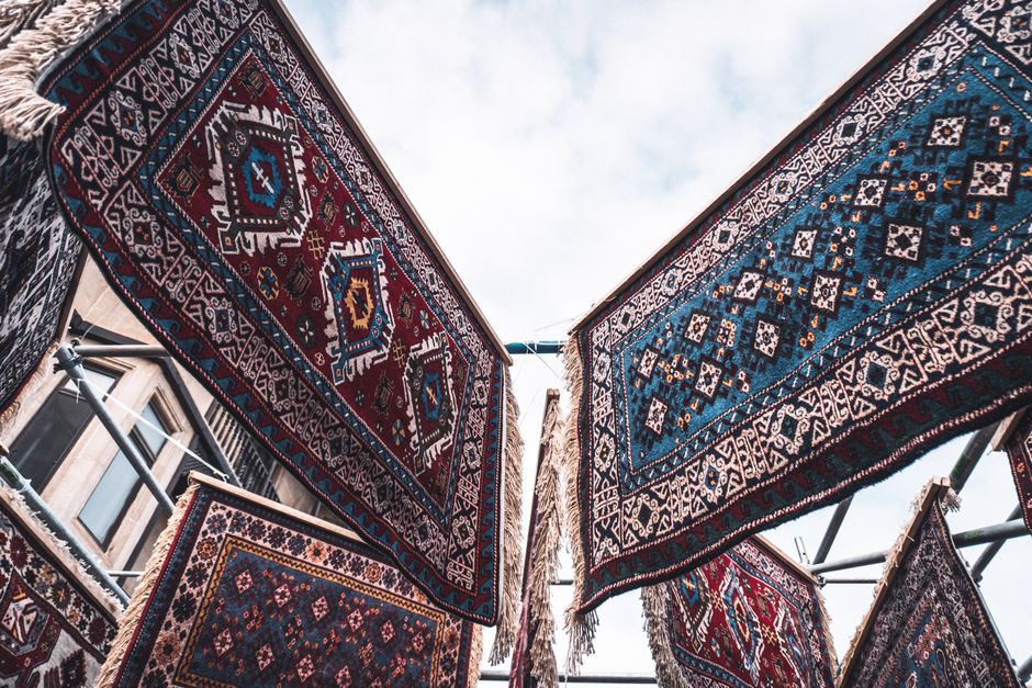 Close-up view of several vibrantly patterned oriental rugs hanging outdoors on a clothesline, showcasing intricate geometric motifs and a mix of red, blue, and beige colors. The rugs are suspended between building facades with visible windows beneath, under a partly cloudy sky. The scene reflects a traditional textile display, with the rugs appearing clean and well-maintained, highlighting detailed craftsmanship. This setting illustrates surface cleaning and maintenance of decorative textiles, aligning with domestic or commercial carpet and rug deep cleaning guidance by Carpet Cleaners Brixton, based on the Carpet cleaning Brixton Market traders SW9 guide.