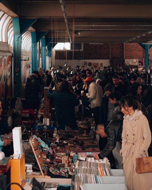 A crowded indoor market with high ceilings and a metal structure, illuminated by natural light through windows and possibly some artificial lighting. The space is filled with numerous stalls covered with various items, such as books, clothing, and accessories, laid out on tables and shelves. The floor appears clean and well-maintained, with visitors browsing tightly packed merchandise. The walls are decorated with minimalistic art and banners, reflecting a lively shopping environment. The overall cleanliness suggests regular maintenance by professional cleaning services like Carpet Cleaners Brixton, ensuring a hygienic space for traders and shoppers in the Brixton Market area. Visible in the background are traders and customers engaged in browsing and purchasing activities, with an atmosphere of bustling commerce and community engagement.